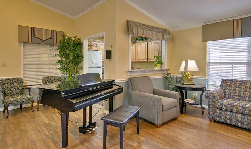 Bright communal living room with a grand piano, upholstered armchairs, a patterned sofa, side table and lamp.