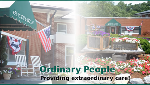 Exterior view of Woodhaven Nursing Home showing a brick building with a green awning labeled Woodhaven, American flags, white rocking chairs on a porch, and a garden area with colorful flowers and wooden planters.