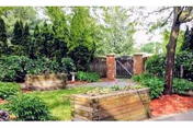 Outdoor garden area with raised wooden planter boxes filled with plants, surrounded by trees and shrubs. There is a wooden fence with a gate in the background and a paved walkway leading through the garden.