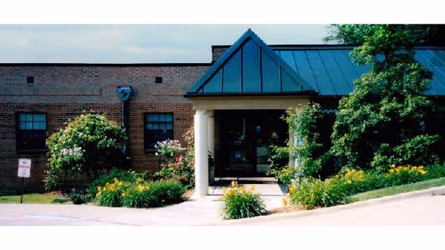 Front entrance of a brick building with a small portico, columns, metal roof, and landscaped flower beds.