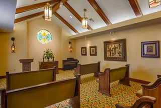 Interior chapel-like room with wooden pews, stained glass window, potted plant, and ceiling beams.