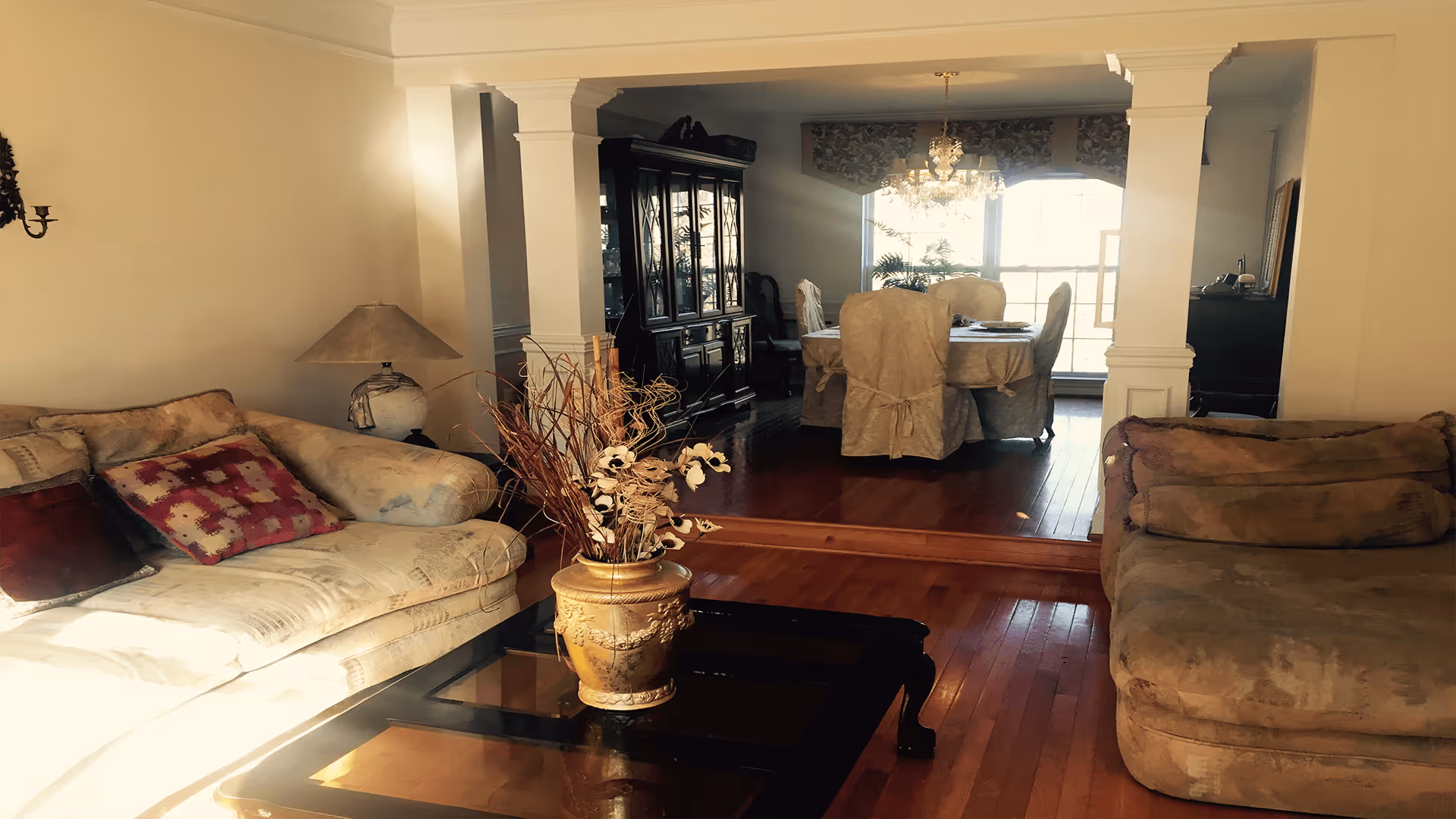 A cozy living room with two beige sofas adorned with cushions, a glass-top coffee table with a decorative vase holding dried flowers, and a lamp on a side table. The room opens into a dining area with a wooden floor, a dining table covered with a cloth, and chairs with slipcovers. A large window in the dining area lets in natural light.