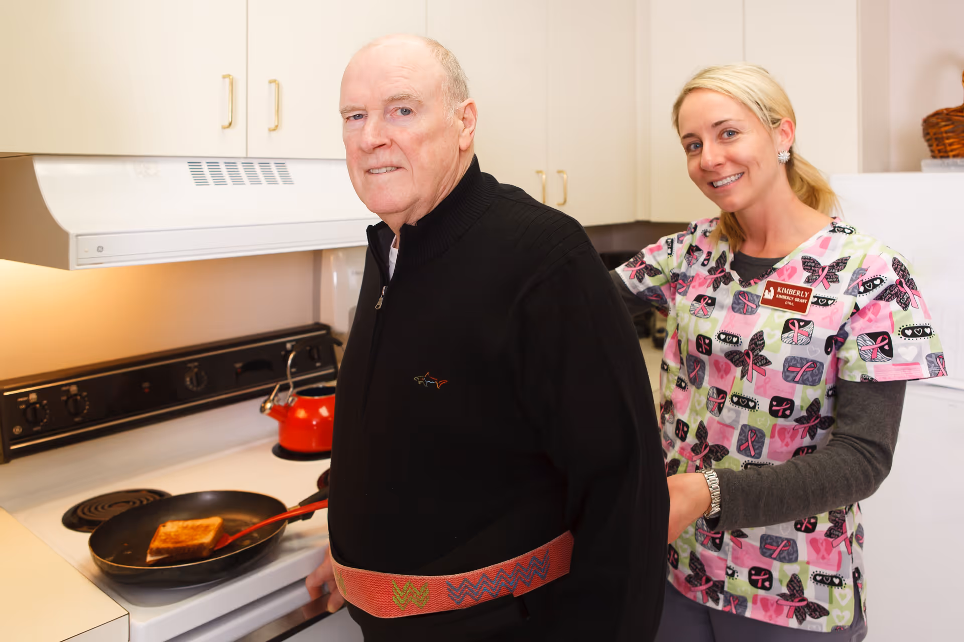 An elderly man is standing in a kitchen cooking a grilled sandwich on a stove while a female caregiver, wearing a colorful patterned scrub top and a name tag that reads Kimberly, assists him by holding a gait belt around his waist. The kitchen has white cabinets, a red kettle on the stove, and a white refrigerator in the background.