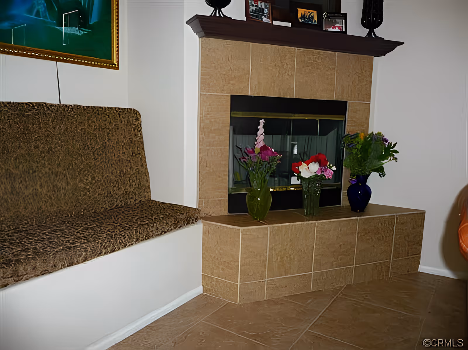 A cozy indoor seating area featuring a built-in bench with a textured brown cushion next to a tiled fireplace. The fireplace mantel holds framed pictures and decorative items, while the hearth displays three vases with colorful flowers. A framed painting hangs on the wall above the bench.