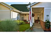 Covered front entrance of the retirement community with an American flag, walkway, porch seating, and landscaping.