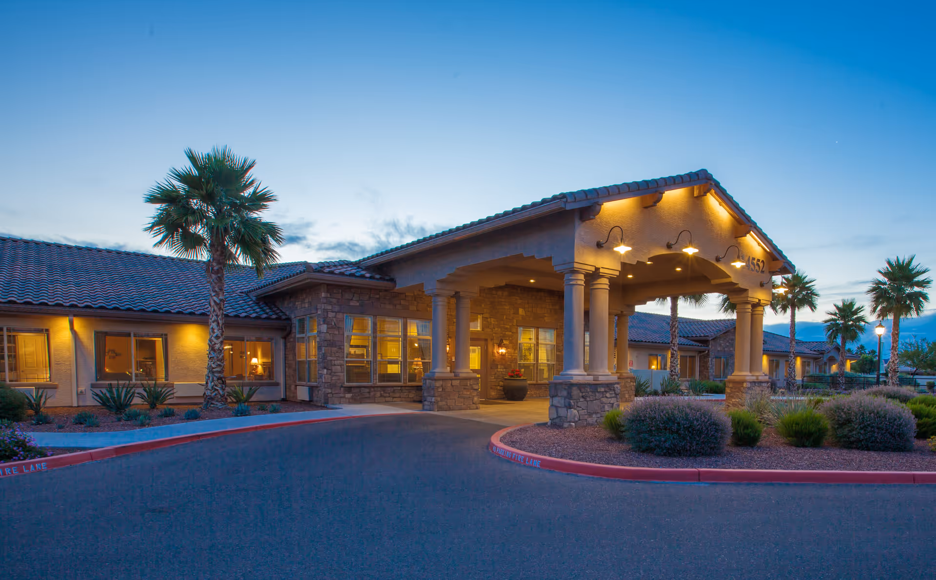 Exterior view of Rock Creek at The Park Memory Care Community building at dusk, featuring a covered entrance with stone pillars, warm lighting, palm trees, and landscaped bushes along the driveway.