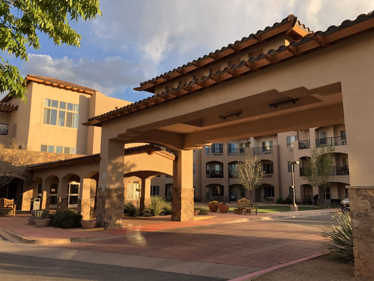 Exterior view of The Rio Grande Gracious Retirement Living facility showing a covered entrance with stone pillars and a tiled roof. The building has multiple balconies and large windows, with some greenery and benches in the courtyard area under a partly cloudy sky.
