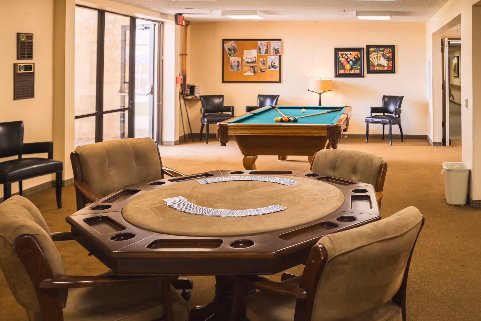 A recreational room with a poker table in the foreground and a pool table in the background. The poker table has playing cards spread out and is surrounded by cushioned chairs. The pool table has pool balls and cues on it. The room has beige walls, carpeted floor, and several black chairs along the walls. There are framed pictures and a bulletin board on the wall, and a lamp providing warm lighting.