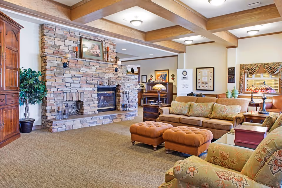 A cozy living room with a large stone fireplace, wooden ceiling beams, and warm lighting. The room features a brown sofa with floral cushions, two matching ottomans, a floral armchair, wooden side tables with decorative lamps, a piano in the background, and framed artwork on the walls.