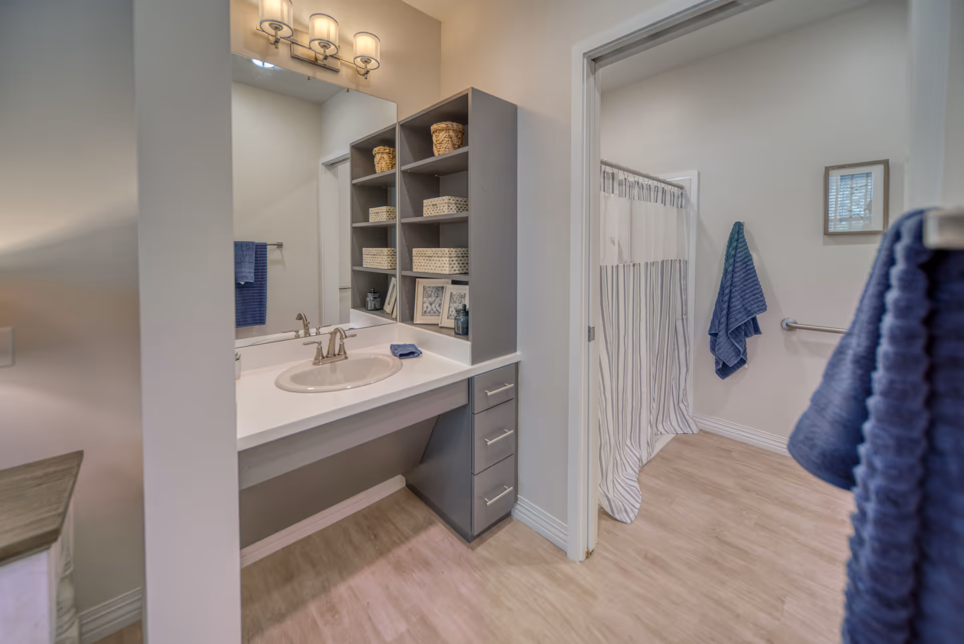 A clean and modern bathroom featuring a white sink with a silver faucet set in a white countertop. Above the sink is a large mirror with a three-light fixture. Next to the sink is a shelving unit with baskets and decorative items. In the background, there is a shower with a white and gray striped curtain, a blue towel hanging on a wall hook, and a grab bar on the wall. The floor is light wood, and the walls are painted a soft neutral color.