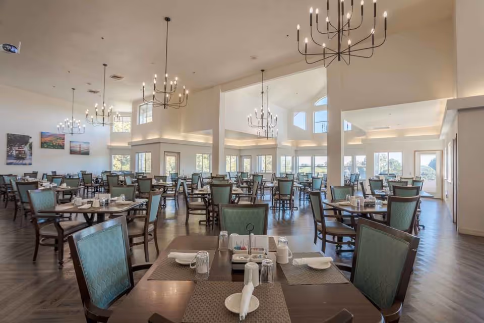Spacious dining room with multiple tables and chairs arranged neatly, each table set with placemats, napkins, glasses, and condiments. The room features large windows allowing natural light to fill the space, high ceilings with modern chandeliers, and light-colored walls adorned with framed artwork.