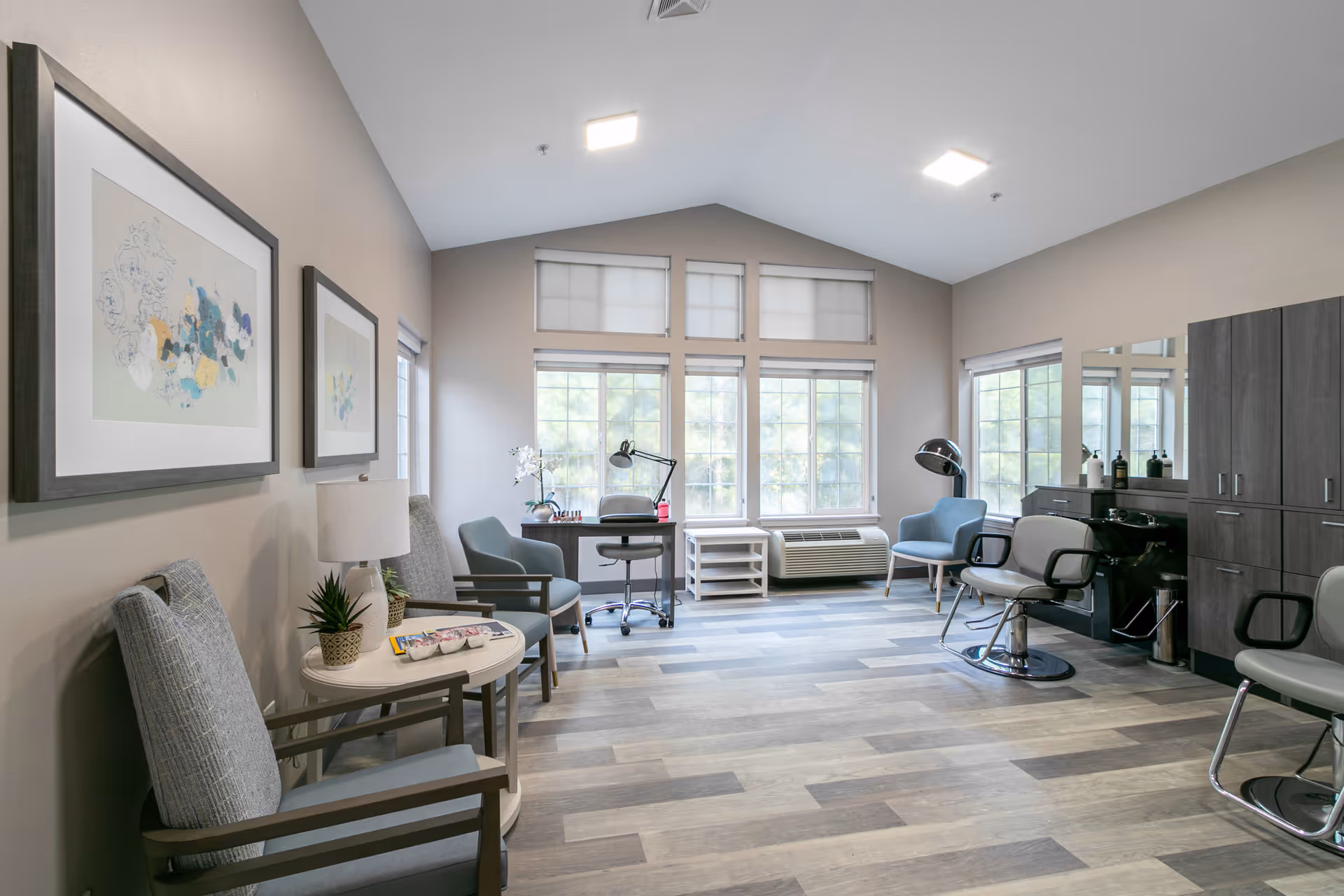 Bright communal salon-style room with styling chairs, a small desk by large windows, seating area and framed wall art.