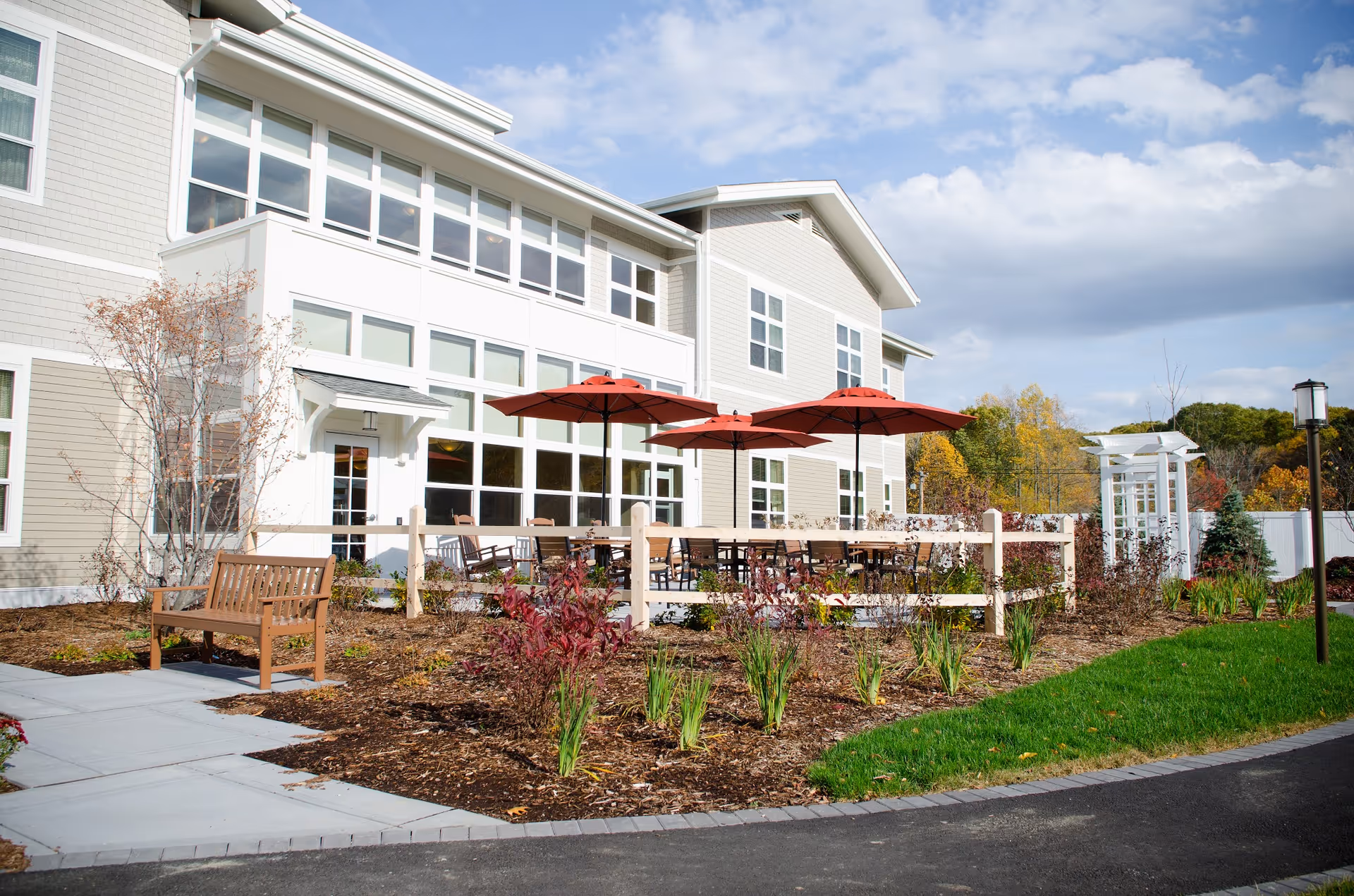 Outdoor patio area of a senior living facility with wooden benches, tables, and red umbrellas. The building has large windows and light-colored siding. There are landscaped garden beds with plants and a paved walkway leading to the patio. The sky is partly cloudy.