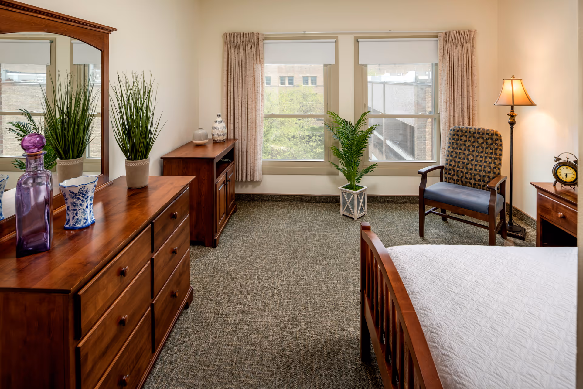 A cozy bedroom with wooden furniture including a dresser with a mirror, a small cabinet, and a bed with a white quilt. There is a patterned armchair next to a floor lamp and a small wooden nightstand with an alarm clock. Two windows with beige curtains let in natural light, and there are green plants placed near the windows and on the dresser.