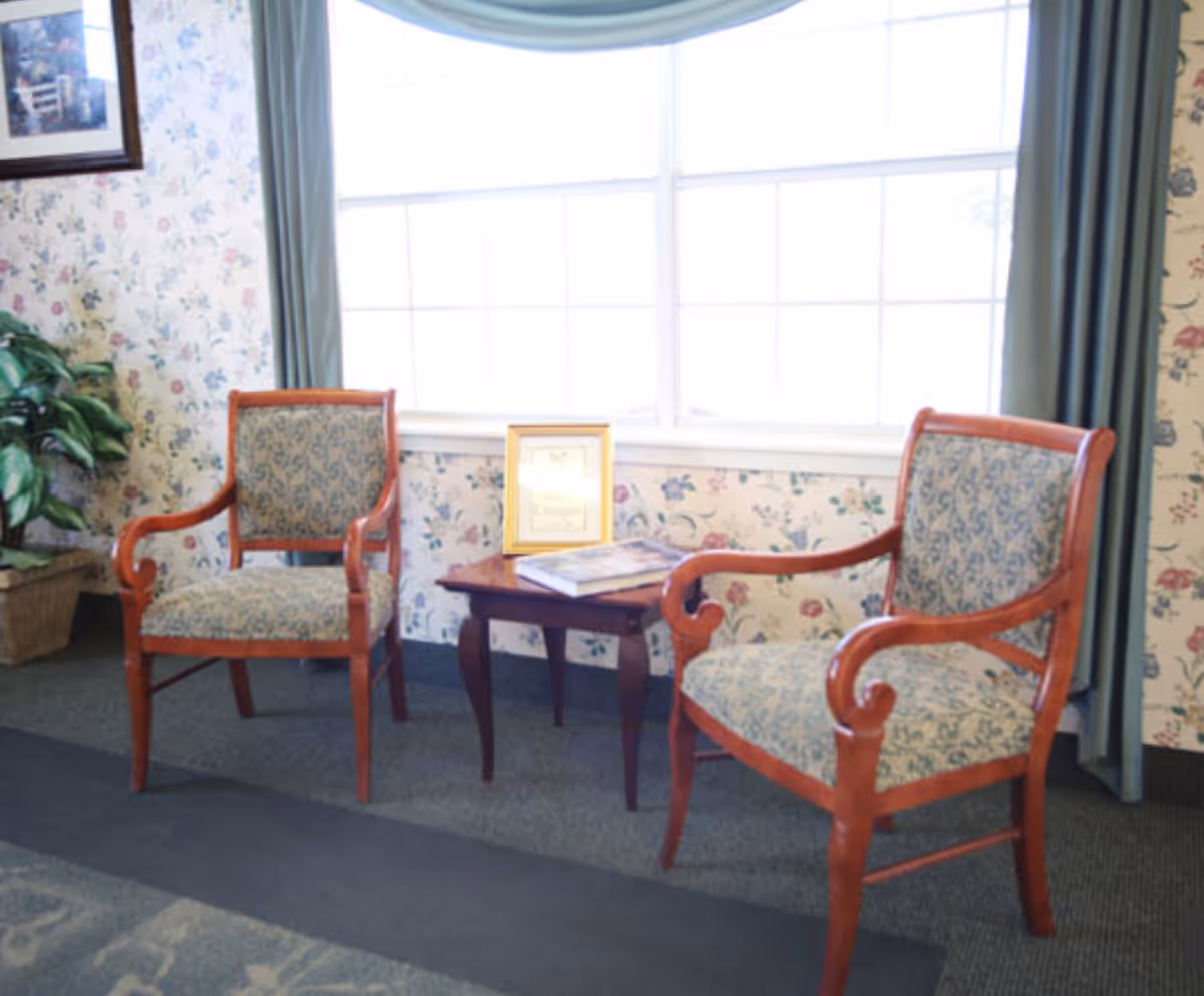 Two upholstered wooden armchairs flanking a small table in front of a large window with floral wallpaper and curtains.