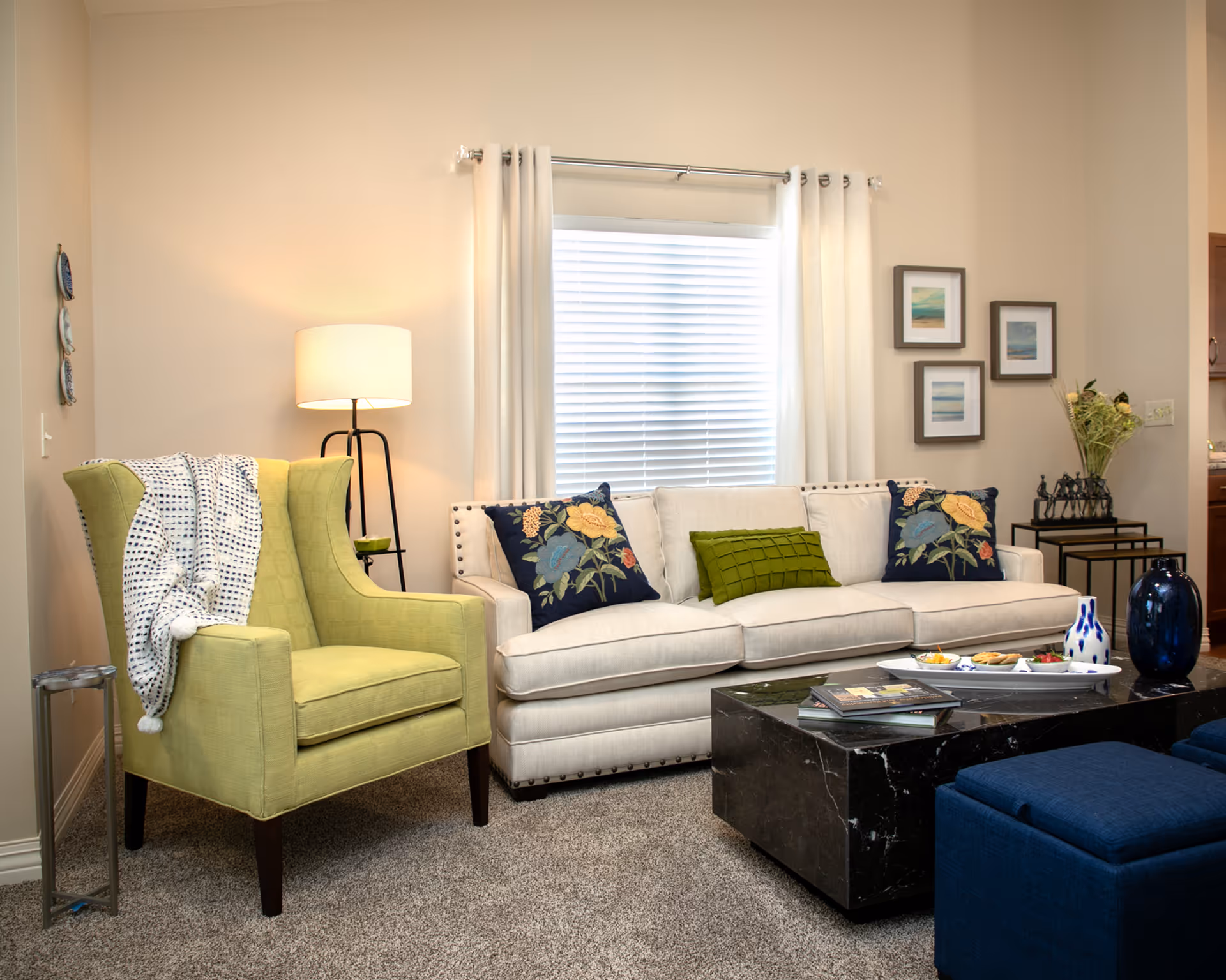 A cozy living room with a light green armchair draped with a white and black patterned throw blanket, a beige sofa with three decorative pillows featuring floral and green designs, a black marble coffee table with books and a tray of snacks, a floor lamp, and framed artwork on the wall next to a window with white curtains and blinds.