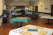 Communal dining room with multiple tables and chairs and a kitchenette/serving area in the background.