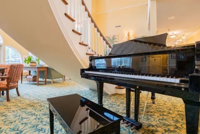 Grand piano and bench in a carpeted common area with a curved staircase and seating in the background.