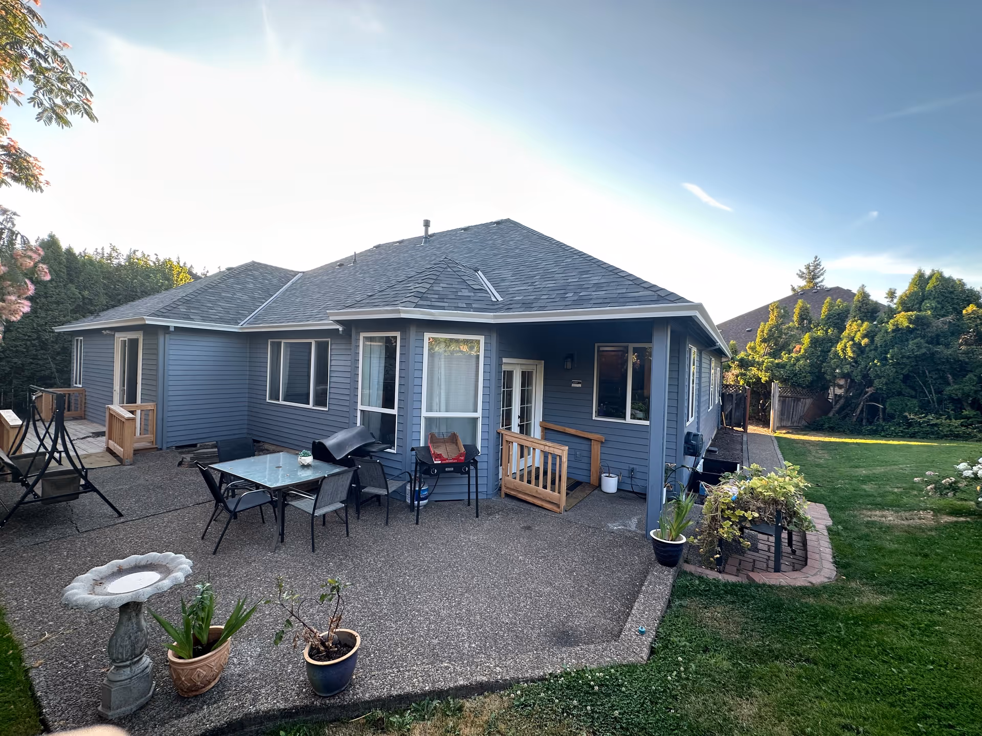 Rear exterior of a single-story blue house with a patio, outdoor furniture, potted plants, and a grassy yard.