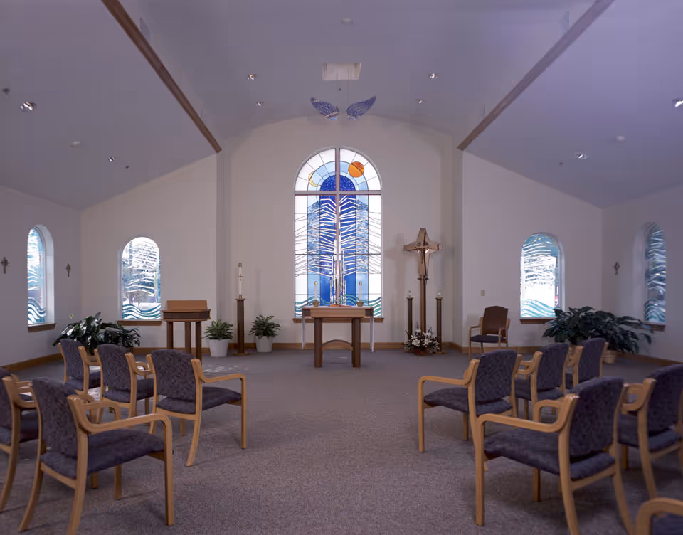 Interior of a chapel with rows of wooden chairs with purple cushions facing an altar. The altar is positioned in front of a large stained glass window depicting abstract designs in blue, white, and orange. There are smaller arched windows on either side with similar stained glass patterns. The room has a high ceiling with recessed lighting and plants placed near the walls.