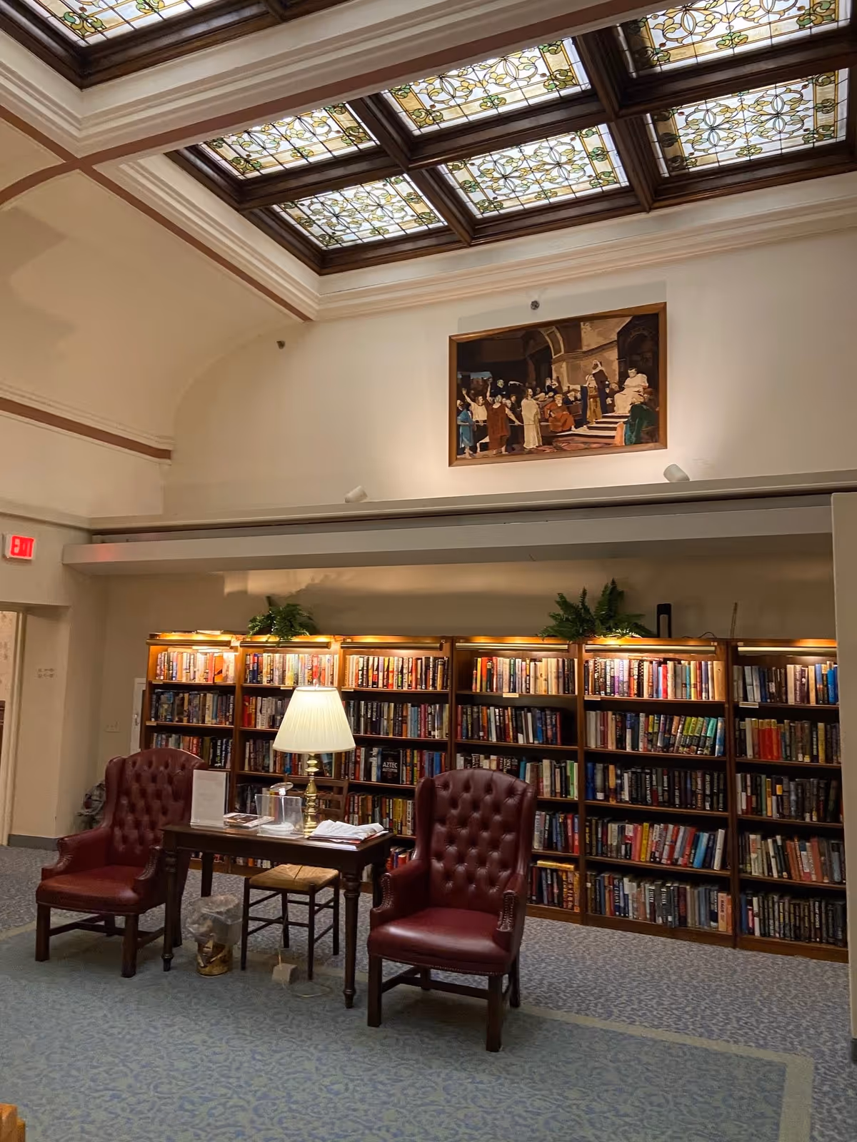 A cozy library room with two red leather armchairs facing a wooden table with a lamp on it. Behind the chairs is a large bookshelf filled with books, topped with green plants and warm lighting. Above the bookshelf is a framed painting depicting a historical or religious scene. The ceiling features decorative stained glass panels.