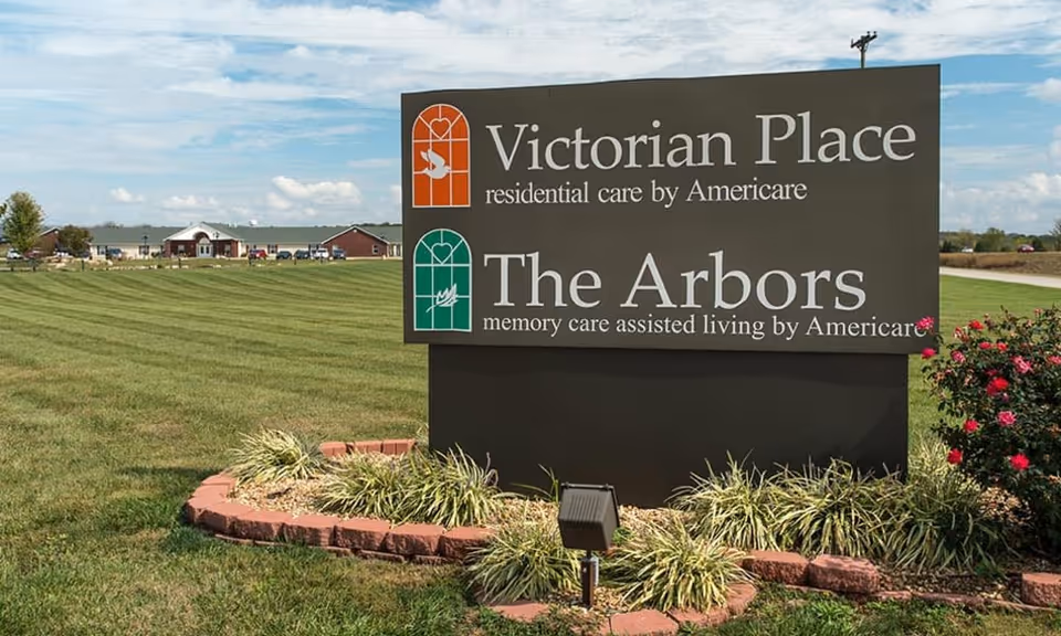 A large roadside sign reading "Victorian Place" and "The Arbors" on a landscaped lawn with the senior living buildings visible in the background.