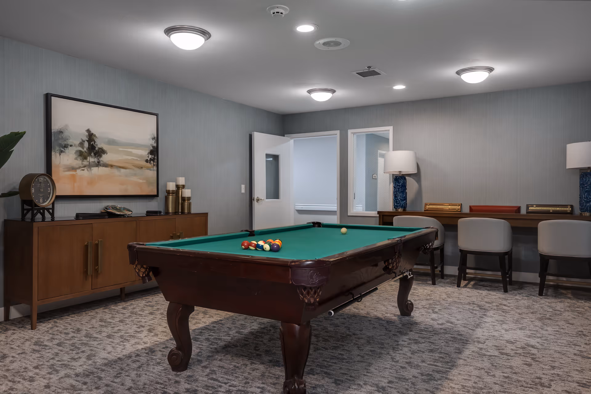 A recreation room featuring a pool table with balls arranged for a game. The room has gray walls, carpeted flooring, and ceiling lights. There is a wooden sideboard with decorative items and a framed landscape painting on the wall. In the background, there is a desk with chairs and two table lamps, and an open door leading to another room.
