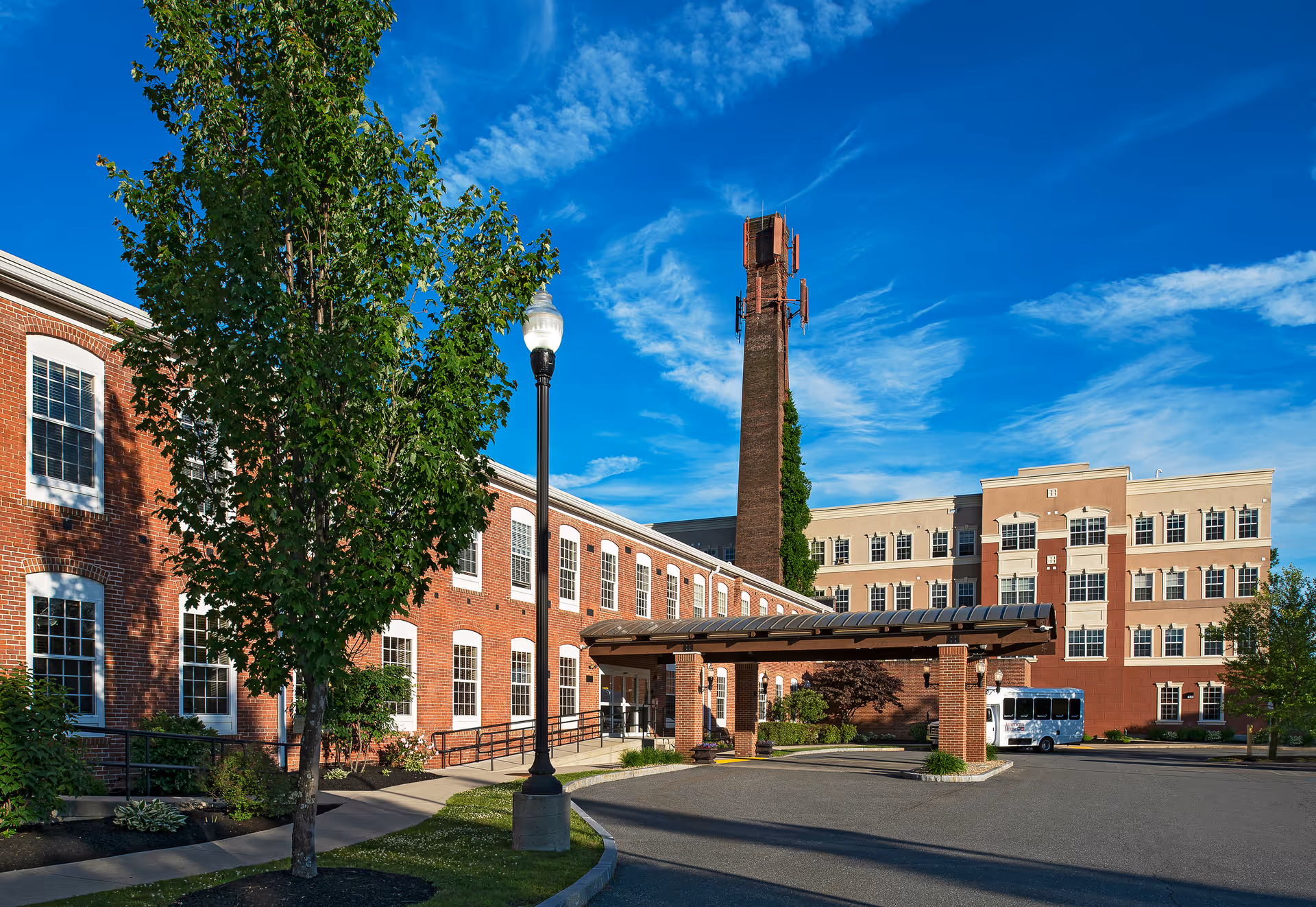 Exterior view of American House Keene facility showing a large brick building with multiple windows, a covered entrance, a tall chimney with antennas, a street lamp, and a small shuttle bus parked near the entrance under a bright blue sky with scattered clouds.