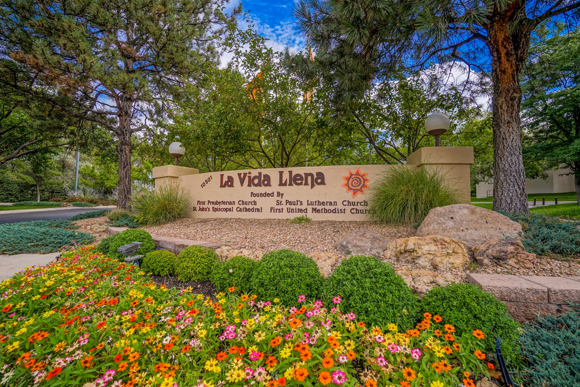 Entrance sign for La Vida Llena Life Plan Retirement Community surrounded by colorful flowers, green bushes, trees, and rocks under a partly cloudy blue sky.