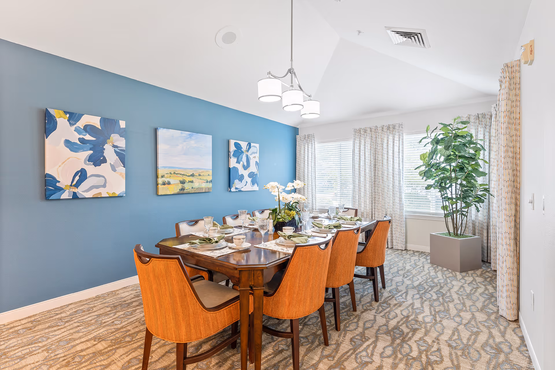A bright dining room with a wooden dining table set for eight people. The chairs have orange upholstery. The room features a blue accent wall with three paintings, large windows with patterned curtains, a potted plant in the corner, and a modern ceiling light fixture.