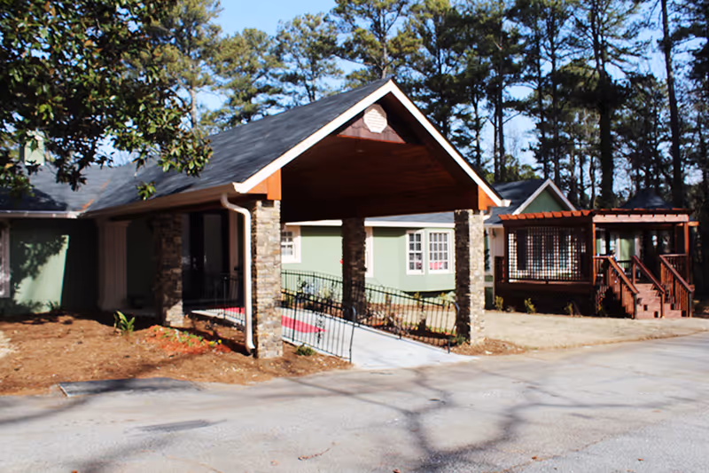 Exterior view of a single-story building with a covered entrance supported by stone pillars, a ramp with handrails, and a wooden deck with stairs. The building is surrounded by trees and a paved driveway.