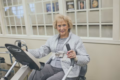 An elderly woman with light hair wearing a gray sweater and pants is sitting on a stationary exercise bike indoors, smiling at the camera. The background shows a wall with windows and framed pictures.