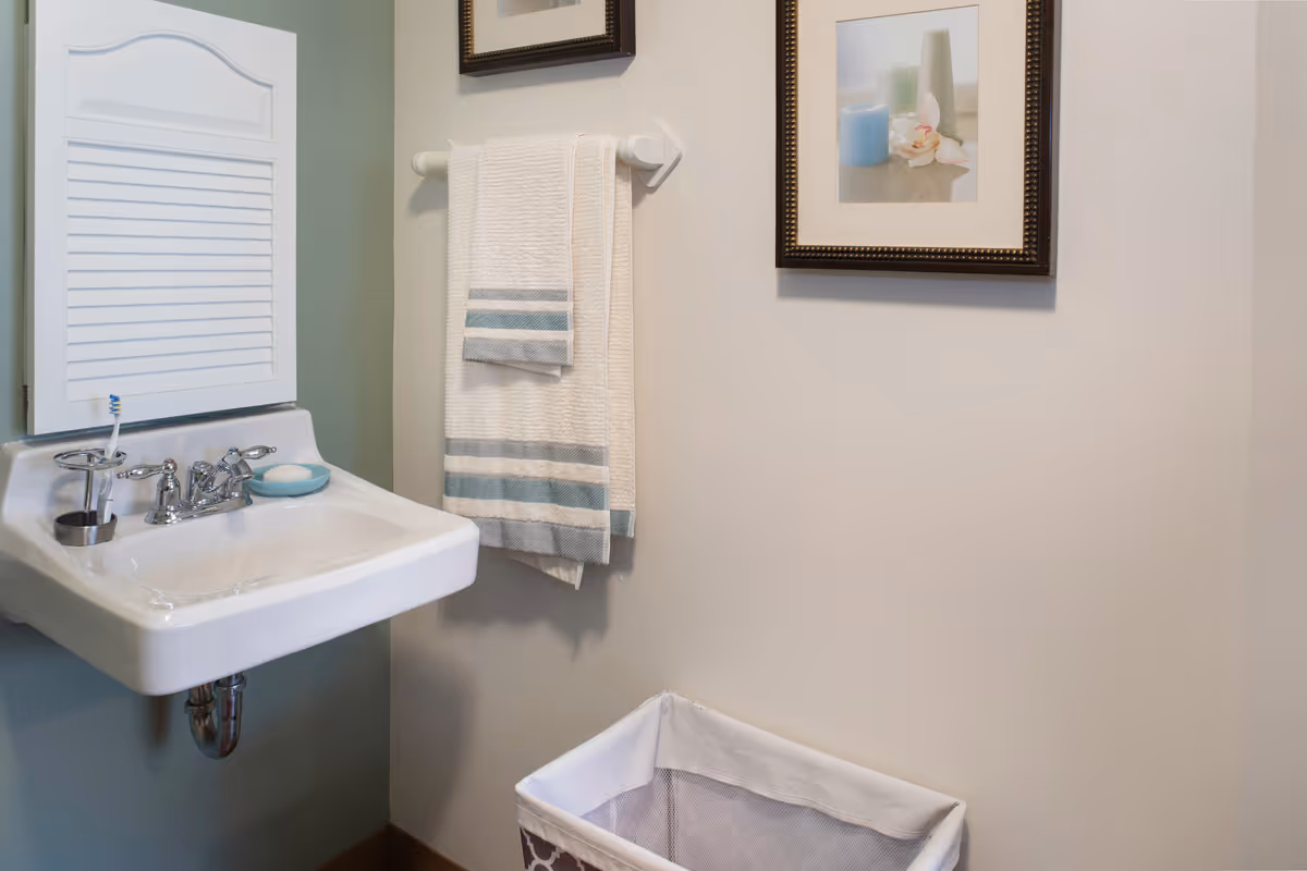 A small bathroom area featuring a white wall-mounted sink with a toothbrush holder and soap dish. Above the sink is a white cabinet with a shutter-style door. To the right, a towel rack holds a white towel with blue stripes. Below the towel rack is a laundry basket, and a framed picture with candles and flowers hangs on the wall.