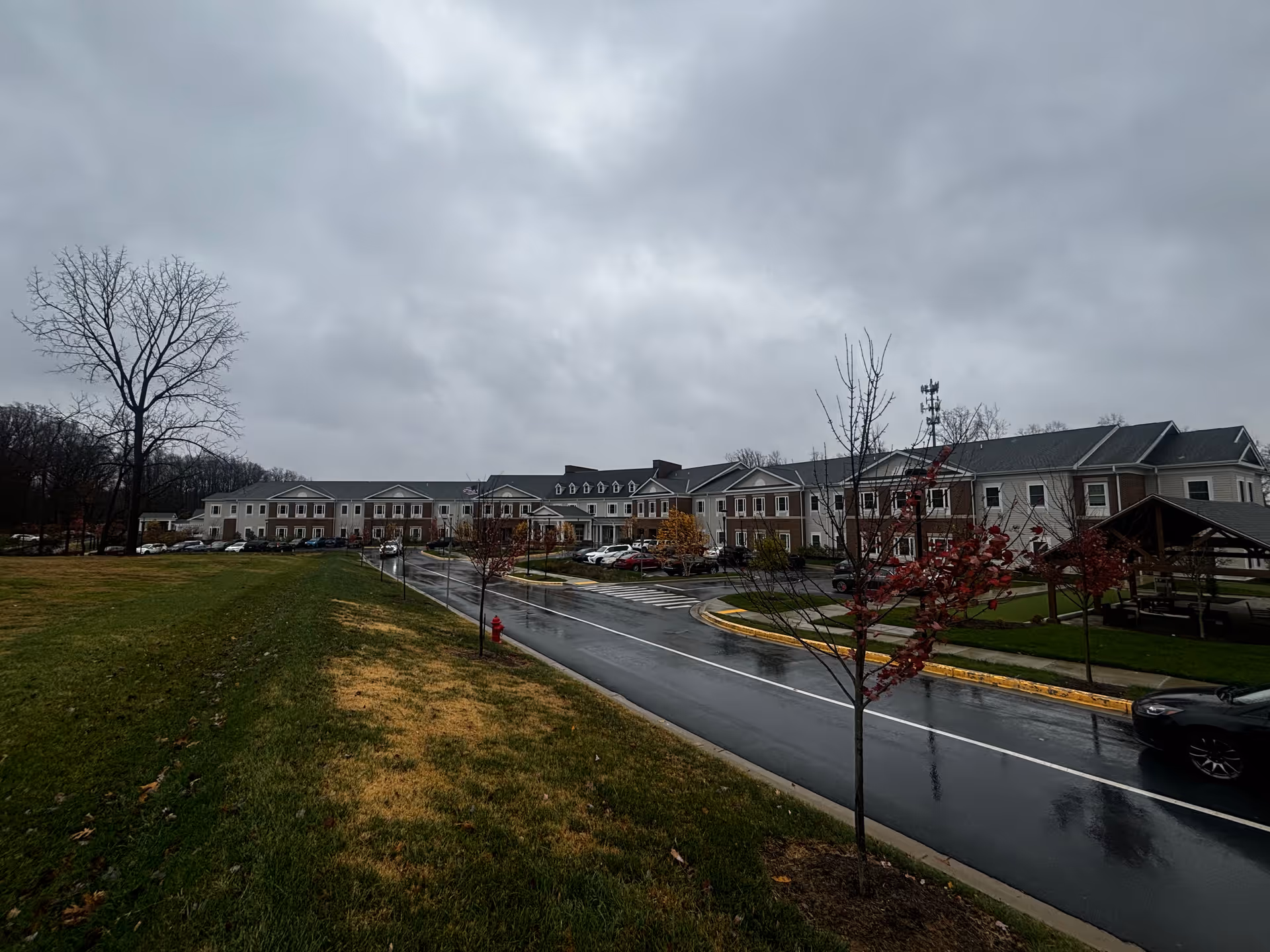 Exterior view of HarborChase of Olney senior living facility on a cloudy day, showing a large two-story building with a parking lot in front, a wet road, some small trees with autumn leaves, and a grassy area.