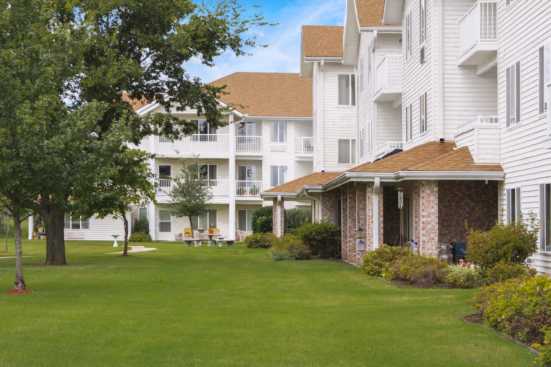 Exterior view of Asher Point Independent Living of Bartlesville showing a multi-story white building with balconies, surrounded by green grass, trees, and shrubs under a partly cloudy sky.