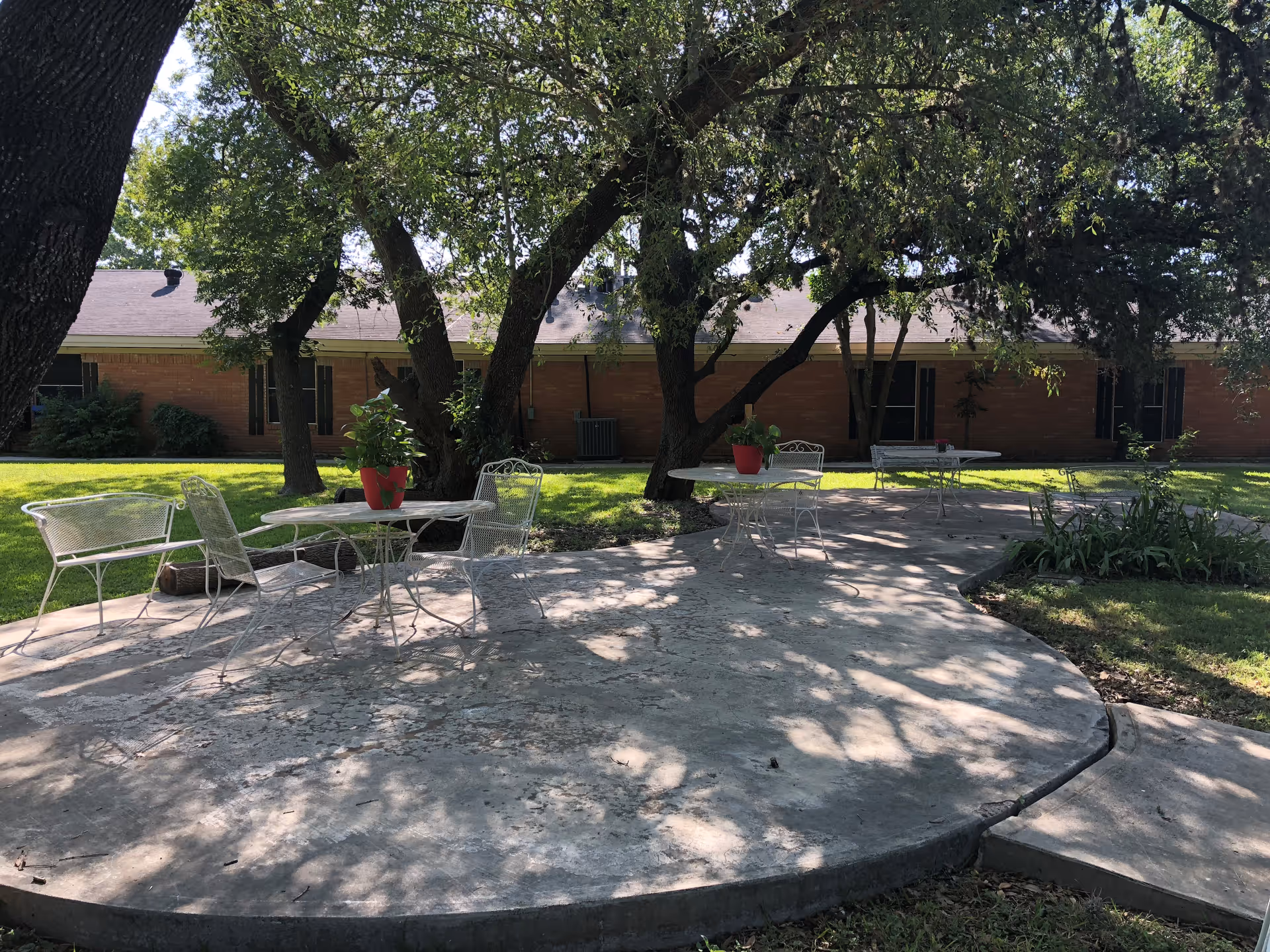 Outdoor patio area with white metal tables and chairs, some with red potted plants on top, surrounded by trees and grass with a brick building in the background.