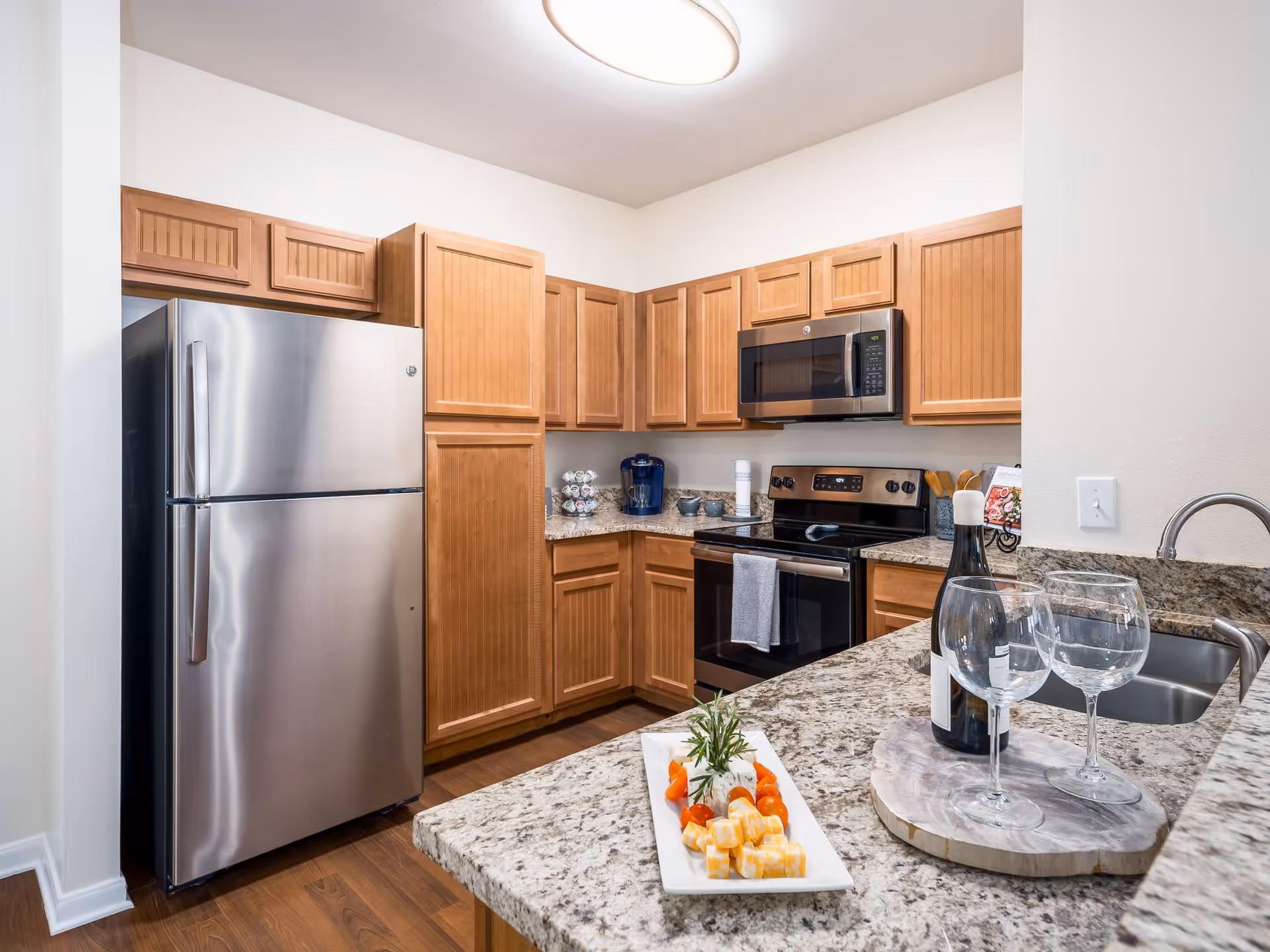 Modern kitchen with wooden cabinets, stainless steel refrigerator, microwave, and stove. Granite countertops with a bottle of wine, two wine glasses, and a plate of cheese and cherry tomatoes are visible. The kitchen has a double sink and a coffee maker on the counter.