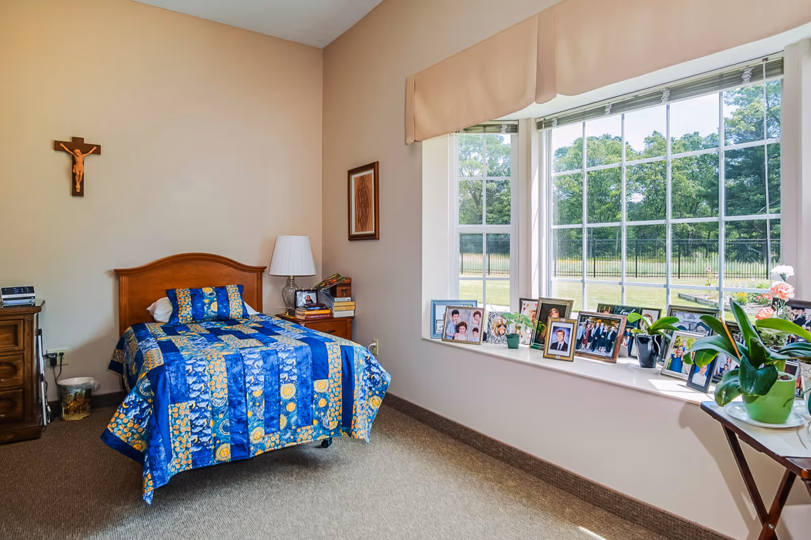 Sunlit bedroom with a twin bed dressed in a blue quilt, a bedside table with a lamp, and a large window sill filled with framed photos and plants.