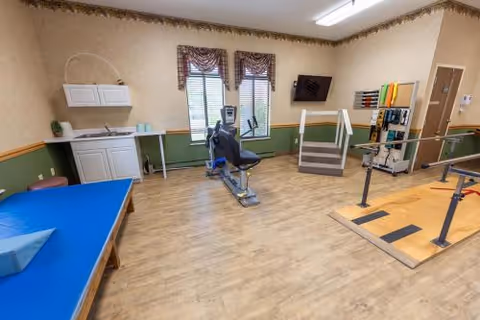 A rehabilitation room with exercise equipment including a stationary bike, parallel bars for walking practice, a blue padded therapy table, and a small set of stairs with handrails. The room has wood flooring, beige walls with green wainscoting, two windows with blinds and valances, a wall-mounted TV, and a small sink with cabinets.