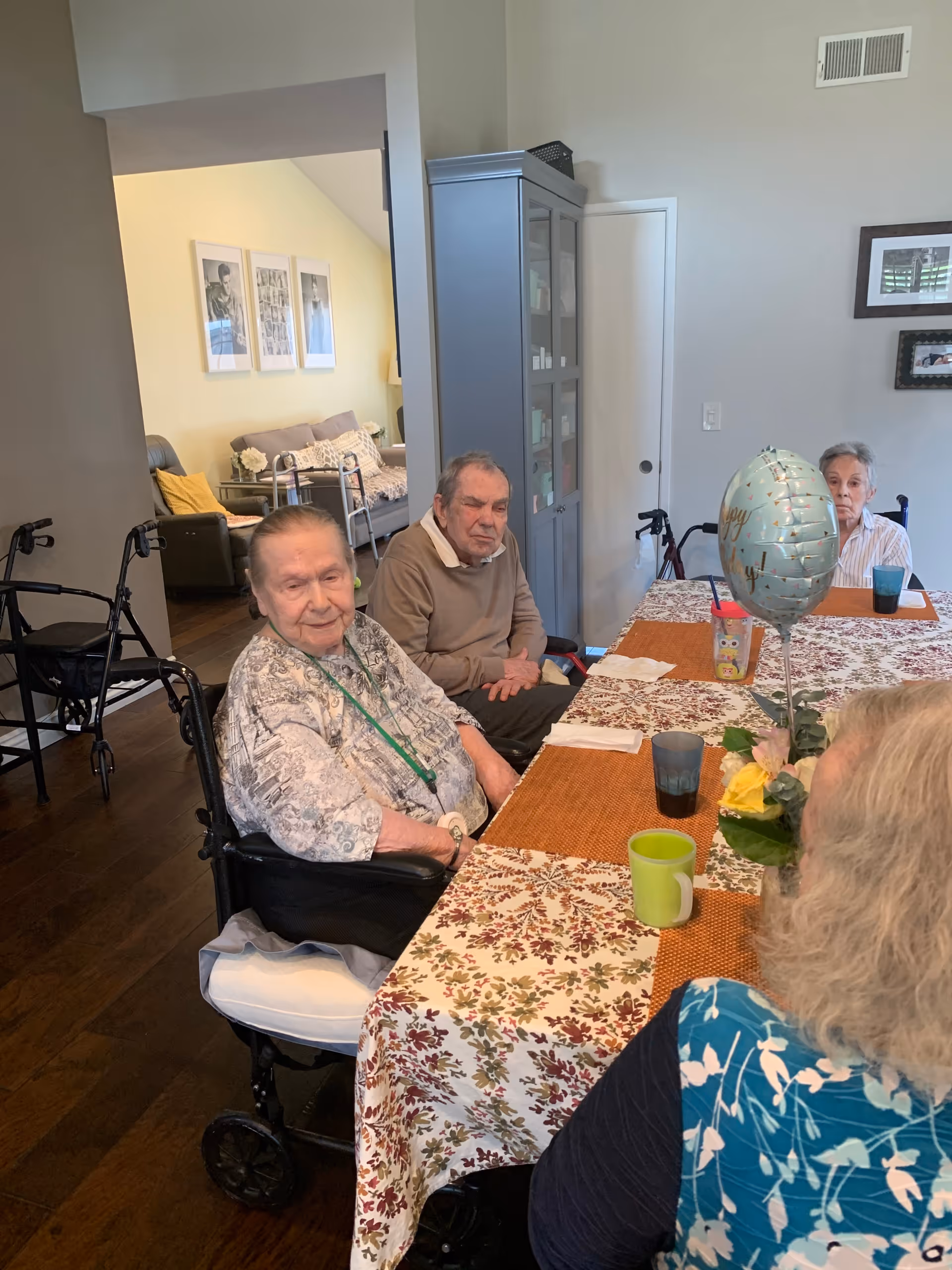 Several elderly residents sit around a decorated dining table in a communal indoor area, with walkers and a balloon visible.