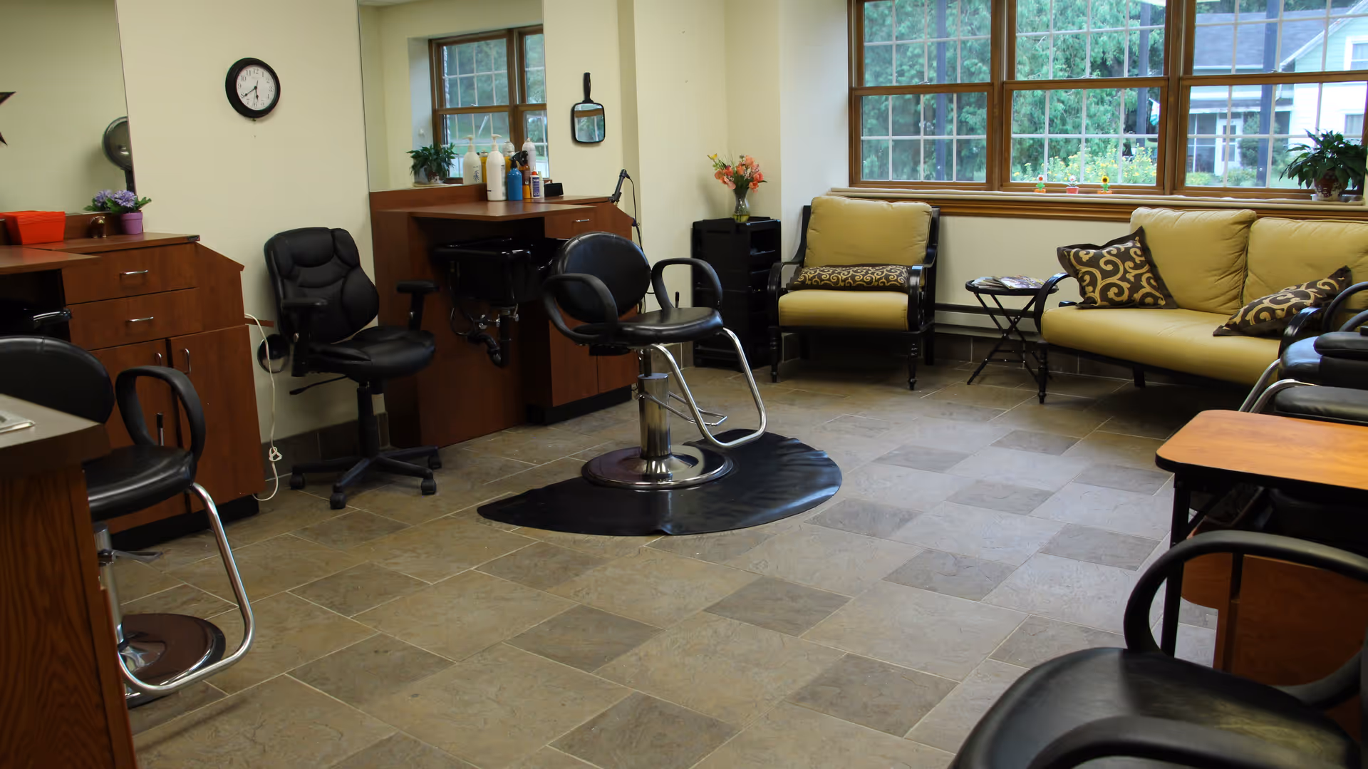 A bright interior salon-style room with a central styling chair, extra salon stations, and cushioned seating by a large window.