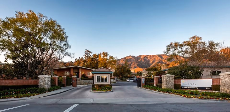 Entrance driveway and guardhouse of the Royal Oaks senior living community with landscaping and mountains in the background.
