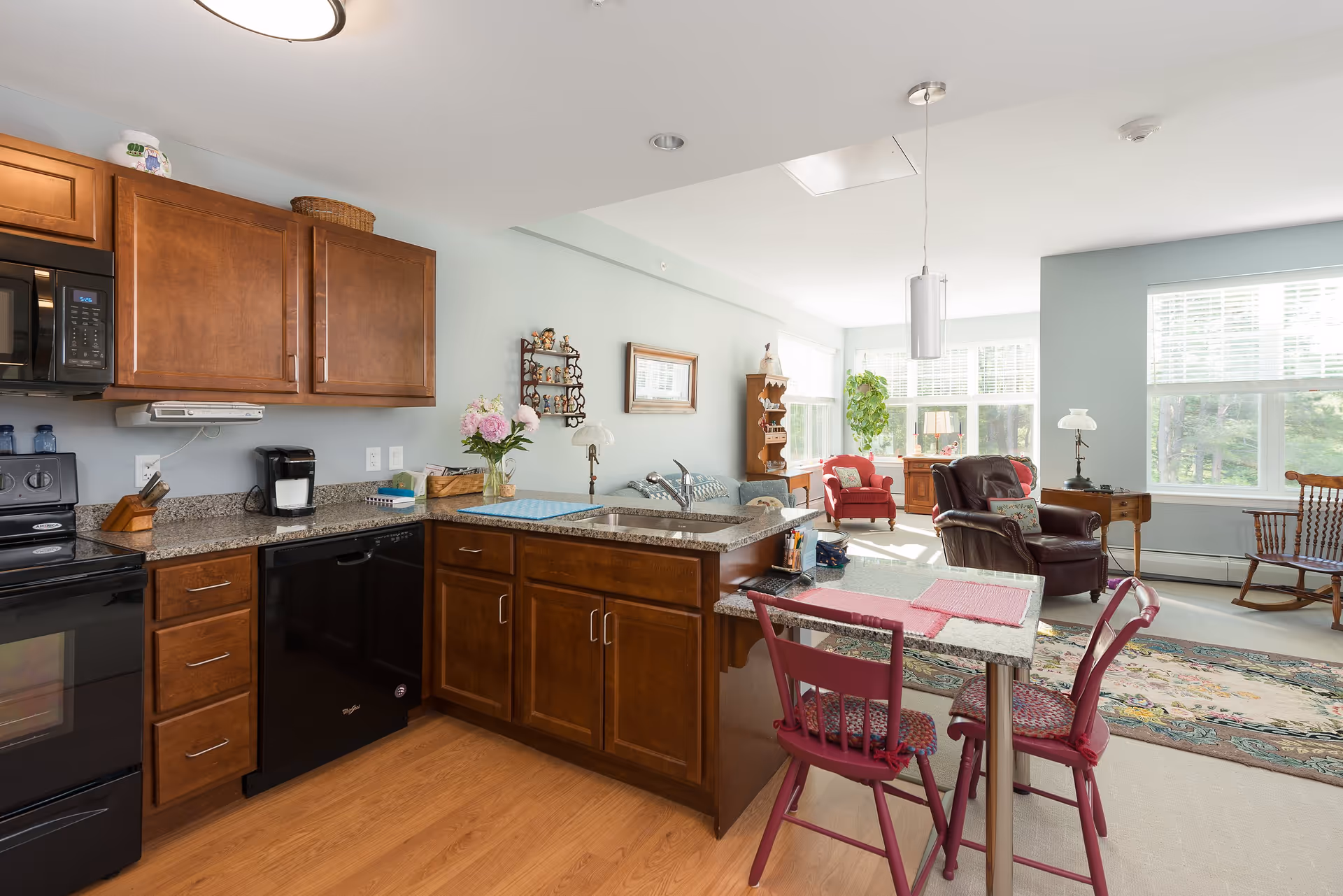 A bright and cozy open-concept living space featuring a kitchen with wooden cabinets, black appliances, and granite countertops. The kitchen island has a small dining table with two red chairs. The living area includes a red armchair, a brown leather recliner, a wooden rocking chair, and large windows letting in natural light. The walls are painted light blue, and there are decorative items such as flowers, a wall shelf, and a framed picture.