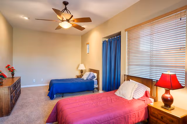 A bedroom with two single beds, one with a red bedspread and the other with a blue bedspread. There is a wooden dresser with red flowers on the left side, a nightstand with a red lamp next to the bed with the red bedspread, and a ceiling fan with a light fixture. The room has beige walls, carpeted floor, and a window with blinds on the right side.