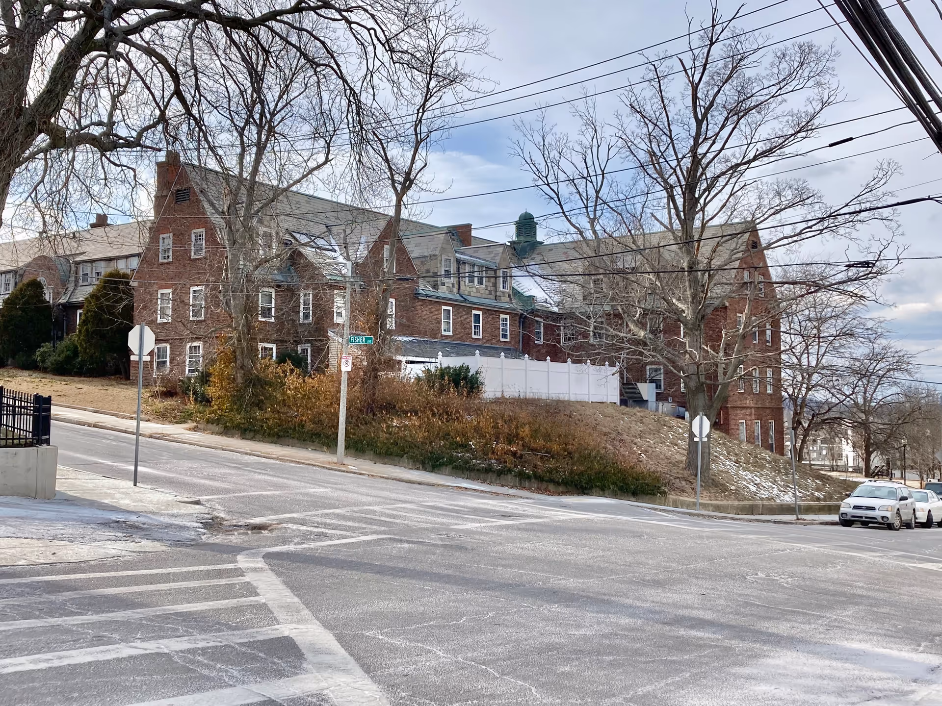 Exterior view of a large, multi-story brick building with a steep roof and multiple windows, situated on a slightly elevated grassy area with leafless trees and a street intersection in the foreground.