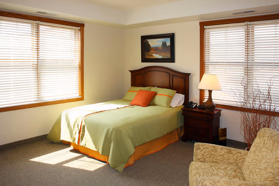 Sunlit bedroom with a wooden bed dressed in green bedding, a nightstand with a lamp, an armchair, and two large windows with blinds.