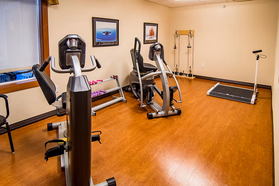 A small exercise room with wooden flooring containing two recumbent exercise bikes, a rack with small dumbbells, a pulley exercise machine, and a scale. The walls are beige with two framed pictures hanging, and there is a window with a brown frame on the left side.