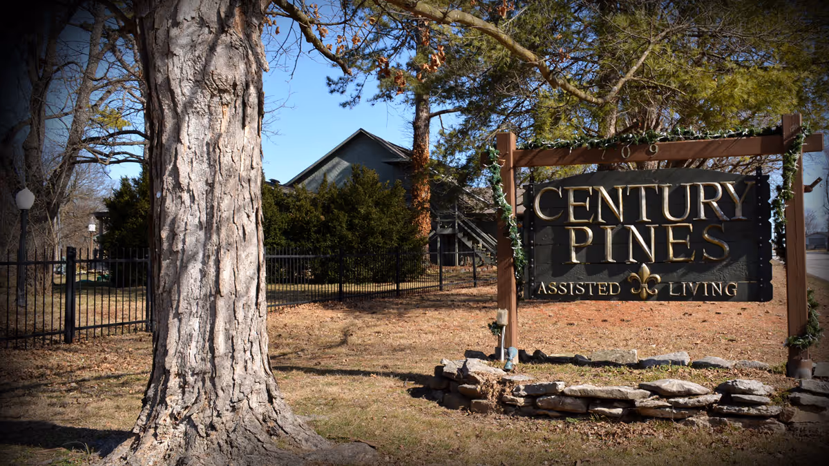 Outdoor view of Century Pines Assisted Living sign surrounded by trees and a stone border, with a building and fence in the background under a clear blue sky.