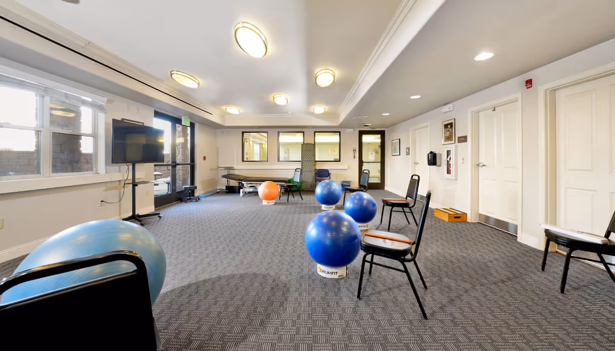 A spacious exercise room with several chairs arranged around large exercise balls on a carpeted floor. The room has white walls, multiple ceiling lights, a TV mounted on a stand near windows and a door leading outside. There is also a treatment table and some framed pictures on the walls.