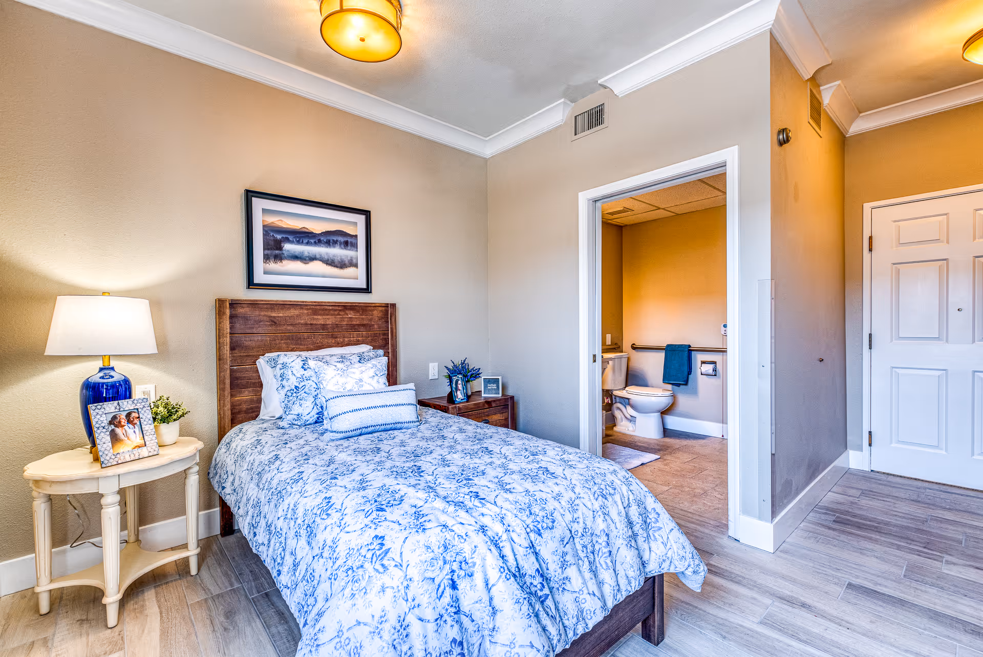 A cozy bedroom with a single bed covered in blue and white floral bedding. There is a wooden headboard, a nightstand with a blue lamp, a framed photo, and a small plant. A framed landscape picture hangs on the wall above the bed. The room has light-colored walls and wood flooring. An open door reveals a bathroom with a toilet, grab bars, and a towel hanging on the wall.